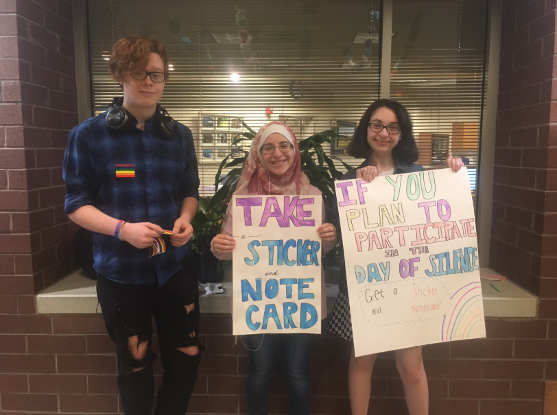 Day of Silence participants Diana Leasburg, Nour Goulmamine and Imke Davis hold up handmade posters explaining to take notecards and stickers.