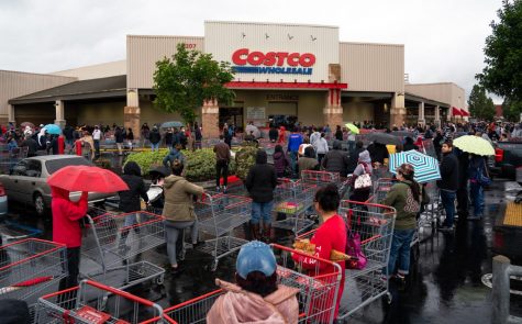 Costco members wait in long lines to enter the store. Signs outside the entrance listed out-of-stock items, with toilet paper and hand sanitizer often at the top of the list.  